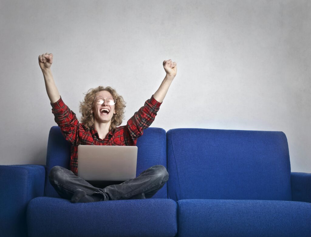 pexels-photo-3770000-3770000 Thrilled individual celebrating success while using a laptop on a blue sofa indoors.